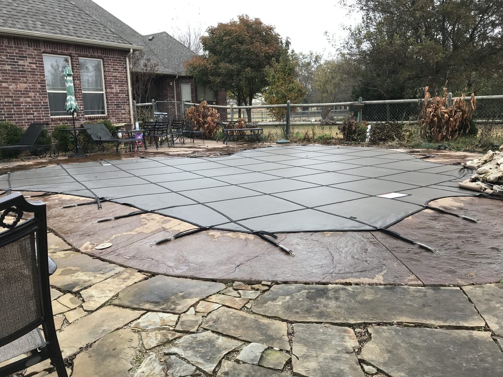 Gray inground pool safety cover on a flagstone and stamped-concrete deck behind a brick home